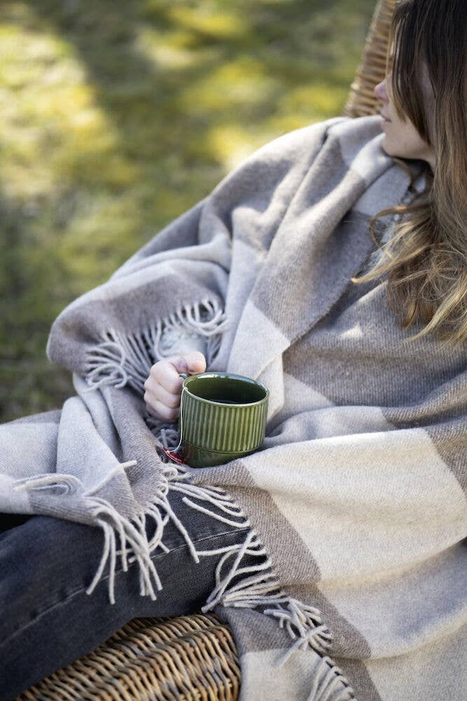 Eine in eine gestreifte Fransendecke gehüllte Person sitzt im Freien auf einem Korbstuhl und hält die Sagaform GmbH Coffee & More Teetasse, 2-tlg. mit abgewandtem Gesicht in der Hand, während das Sonnenlicht durch den Hintergrund scheint.