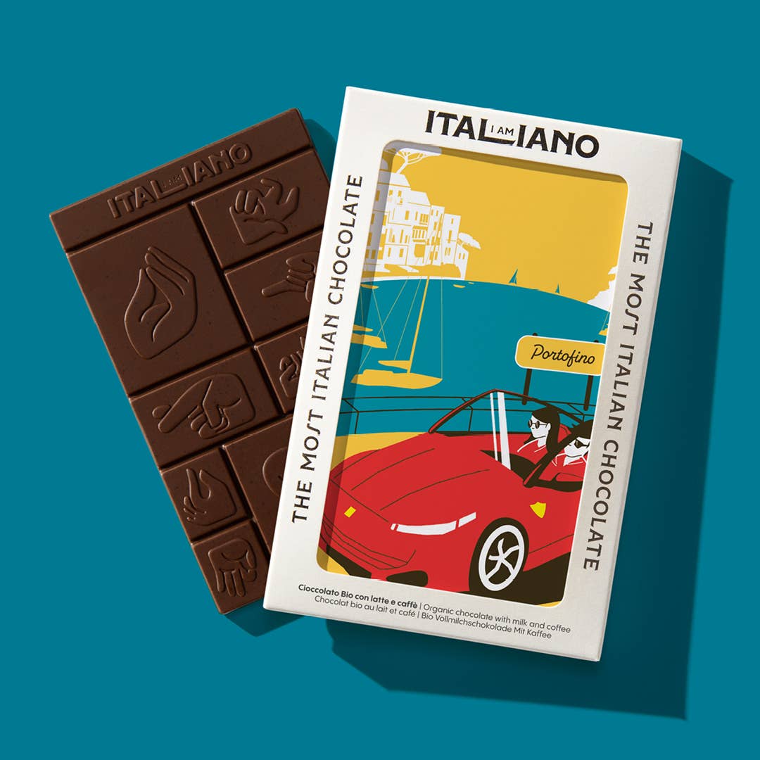 Die I AM ITALIANO - Porto Fino Bio-Schokolade mit Milch und Kaffee, die mit italienischen Handgesten gestaltet ist, wird neben ihrer farbenfrohen Verpackung mit einer illustrierten Frau gezeigt, die ein rotes Cabrio im Küstenort Portofino fährt.