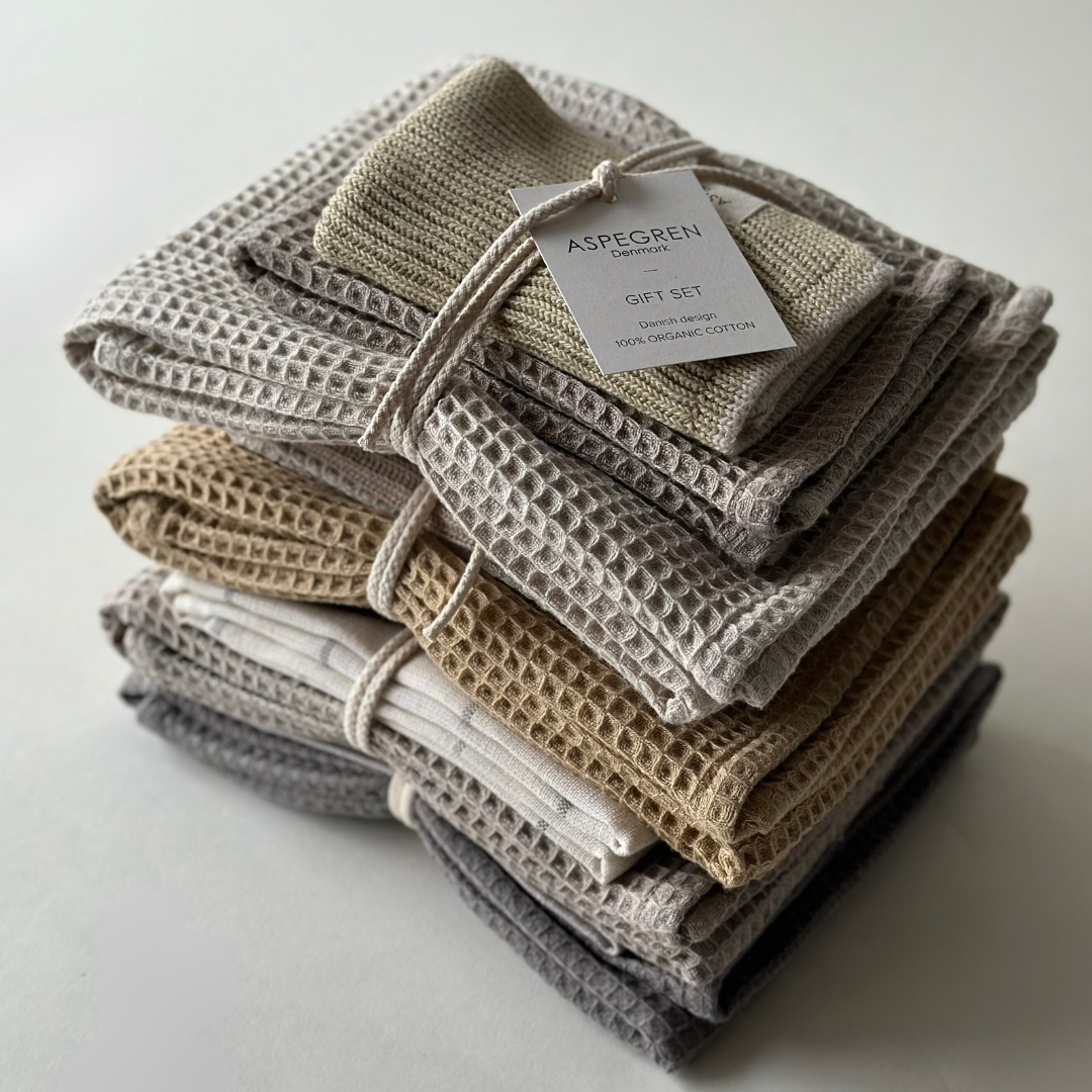 Ein ordentlich gestapeltes Set umweltfreundlicher Handtücher mit Waffelstruktur von Aspegren Denmark – Geschenkset – Lemon Grass in neutralen Farbtönen, mit Schnur zusammengebunden und mit der Aufschrift „100 % Bio-Baumwolle“ versehen, präsentiert auf einer schlichten weißen Oberfläche.