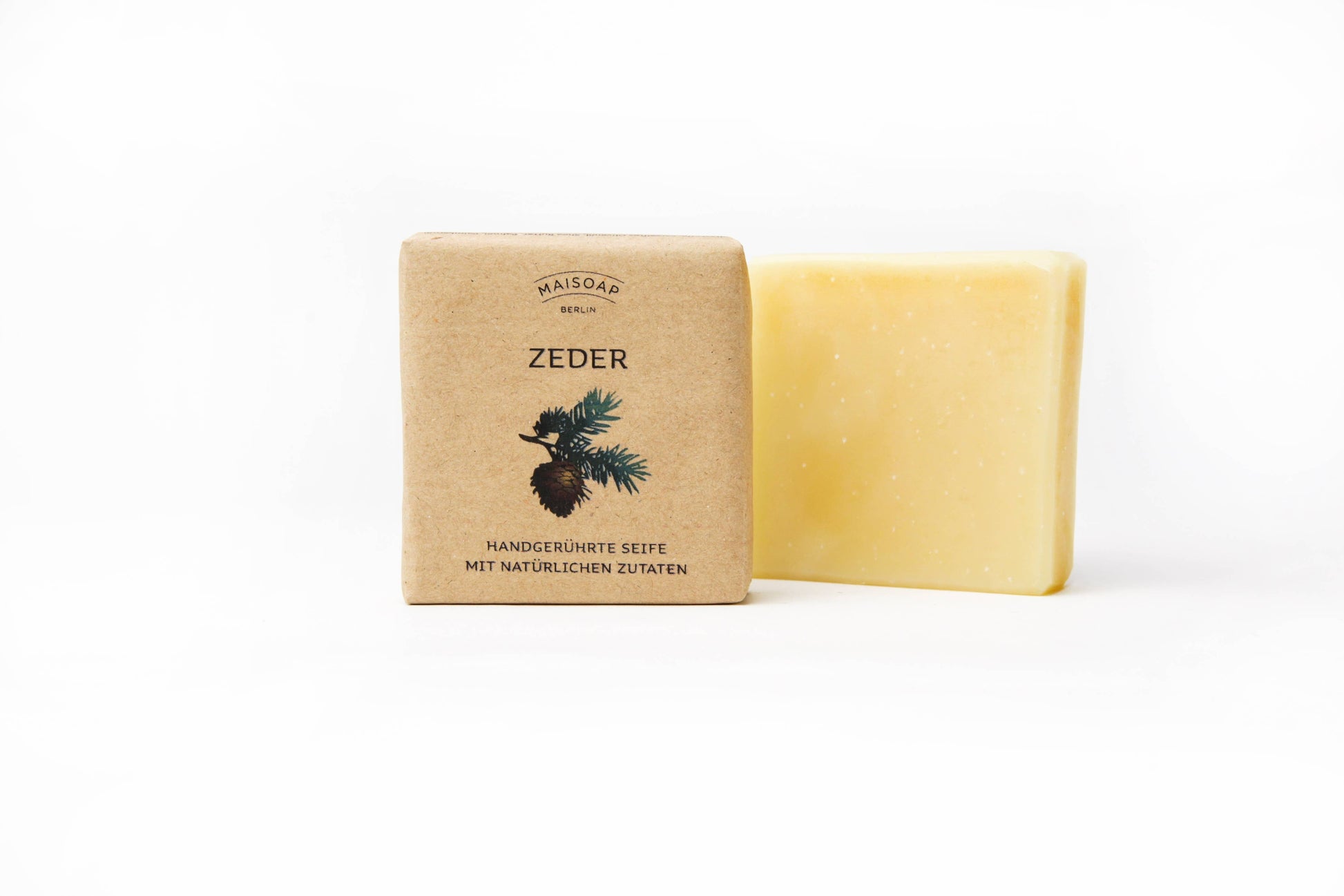 Ein Riegel gelber Zeder Seife von maisoap berlin liegt neben seiner braunen Papierverpackung, auf der ein Tannenzweig und die Aufschrift "Handgerührte Seife mit natürlichen Zutaten" zu sehen sind. Die Seife enthält reichhaltige Sheabutter und einen frischen Zedernduft.