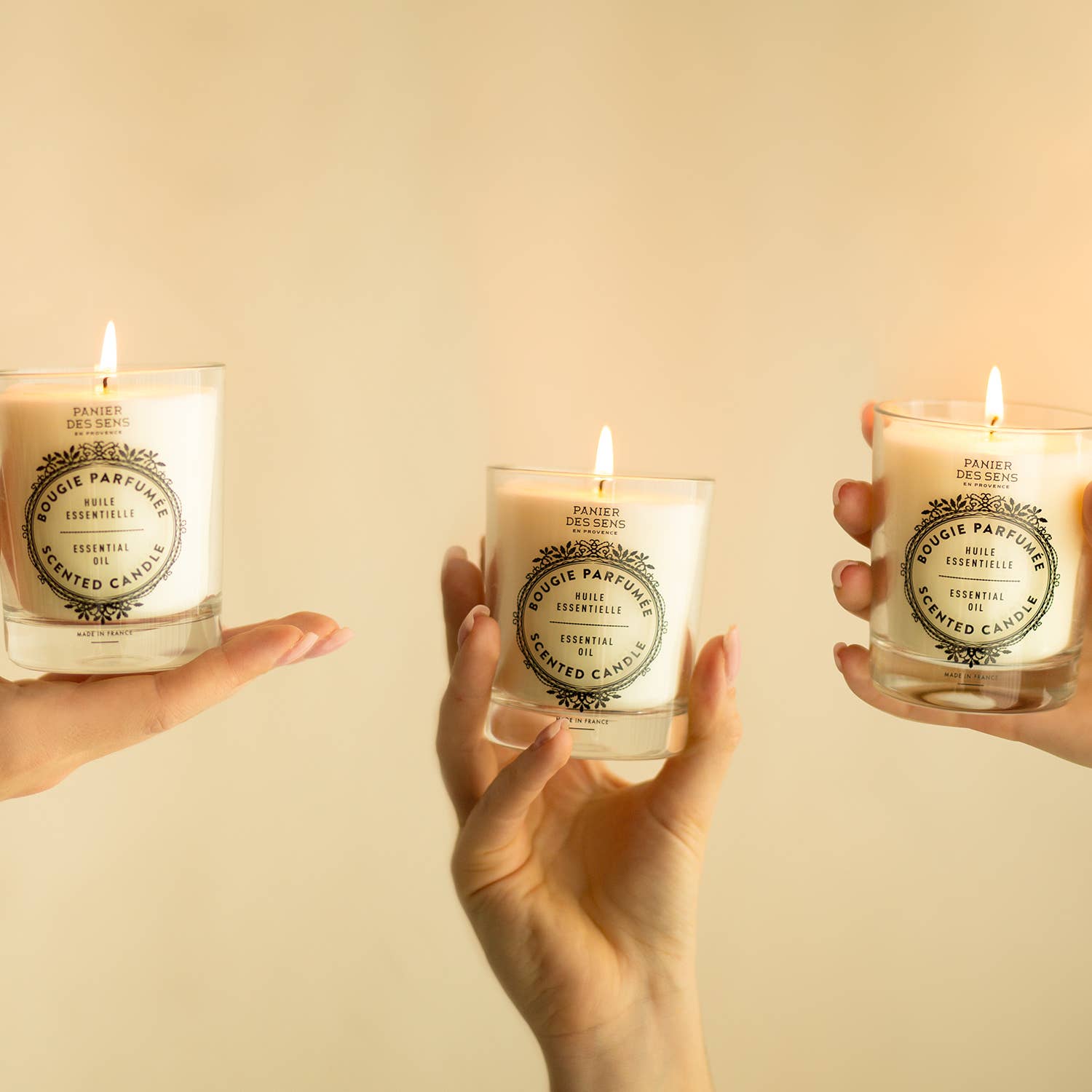Drei Hände halten jeweils eine beleuchtete weiße Panier Des Sens Duftkerze | Provence 180g in einem Glastiegel mit der Aufschrift Bougie Parfumée, mit ätherischen Ölen, vor einem neutralen beigen Hintergrund.