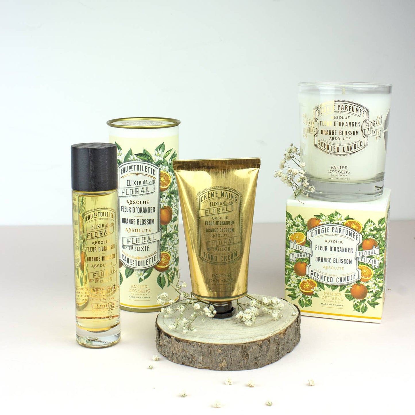 Ein bezauberndes Arrangement von Panier Des Sens-Produkten, darunter die Duftkerze | Orangenblüte 180 g, mit blumen- und orangengeschmückter Verpackung, eingebettet in weiße Blumen auf Holz.