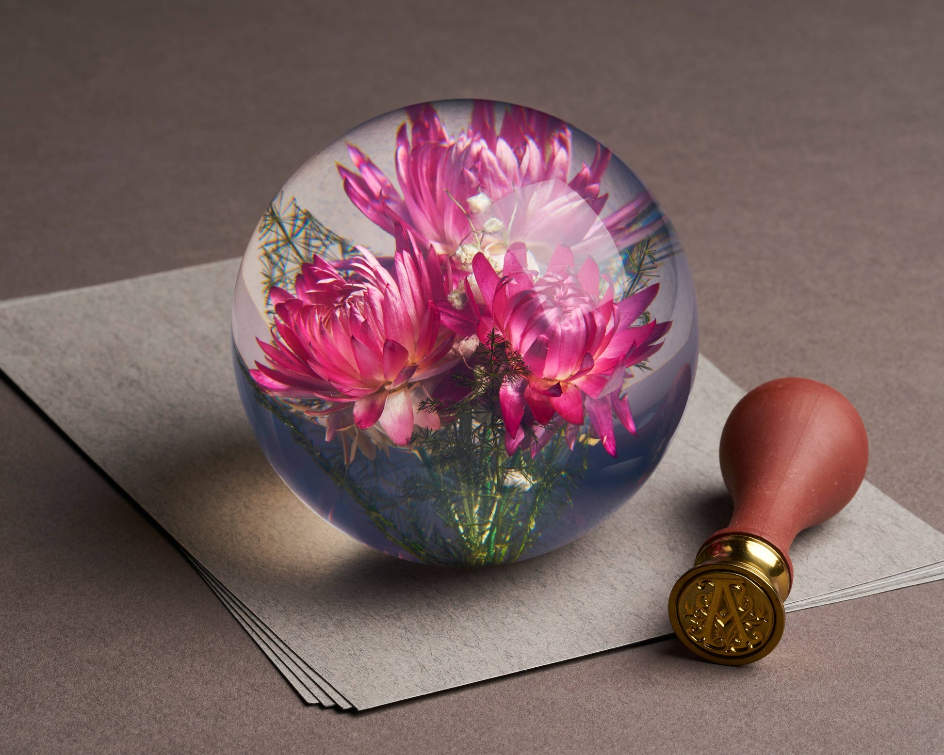 Der Hafod Grange Ltd - Helichrysum Pink Briefbeschwerer, ein Briefbeschwerer aus Glas mit rosa Helichrysum-Blüten von Hafod Grange Ltd, ruht auf gestapeltem grauem Papier neben einem Wachssiegelstempel mit Holzgriff und goldgeprägtem Ende.