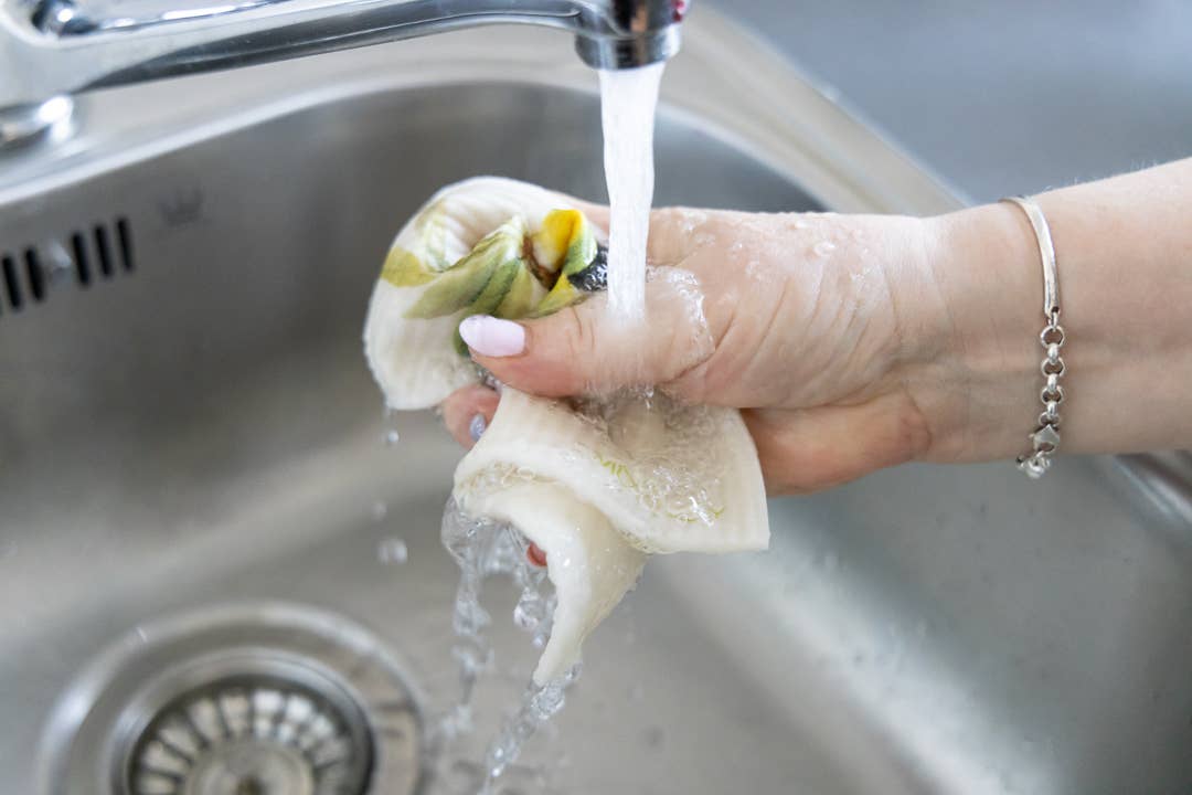 Eine Hand mit einem silbernen Armband spült einen dekorativen Teller unter fließendem Wasser in einer Edelstahlspüle ab, während ein More Joy Schwammtuch Tomaten von More Joy daneben liegt.