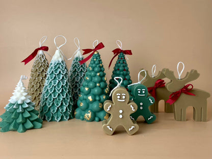 SOULCandle - Weihnachtskerzen von SOULCandle: Handgefertigte Dekokerzen in Form von Weihnachtsbäumen, Lebkuchenmännchen und Rentieren auf beigem Untergrund. Hergestellt mit Dochten aus umweltfreundlichem Sojawachs zum Aufhängen oder Anzünden; einige Bäume sind mit roten Schleifen versehen.