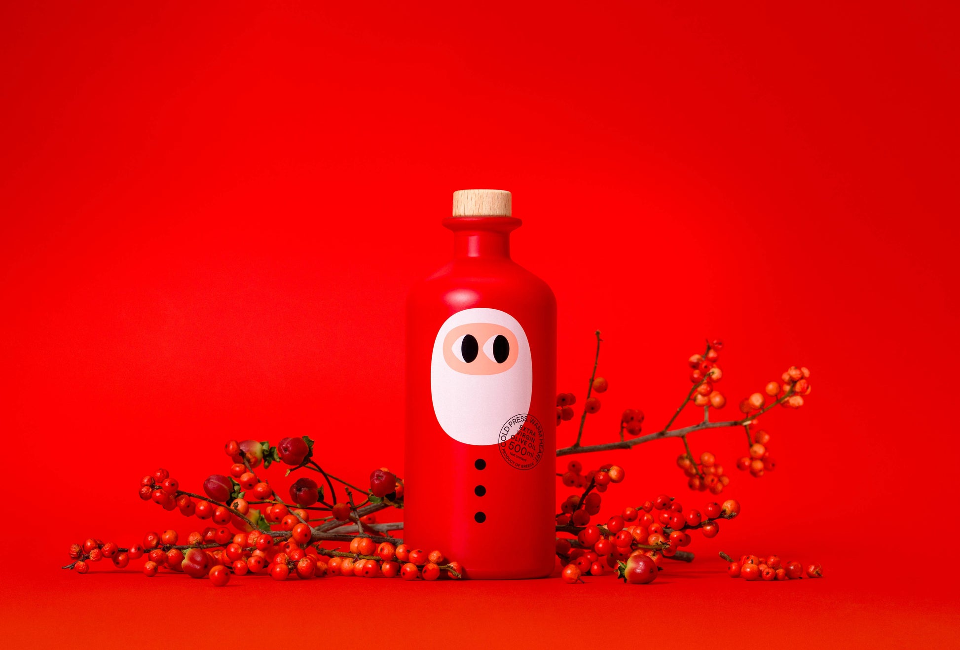 Eine festliche 500 ml Flasche yiayia and friends - Natives Olivenöl extra Weihnachtsedition mit Cartoon-Gesicht und Korkverschluss steht zwischen roten Beeren - ein minimalistisches, limitiertes Design, perfekt als Geschenk für Feinschmecker.