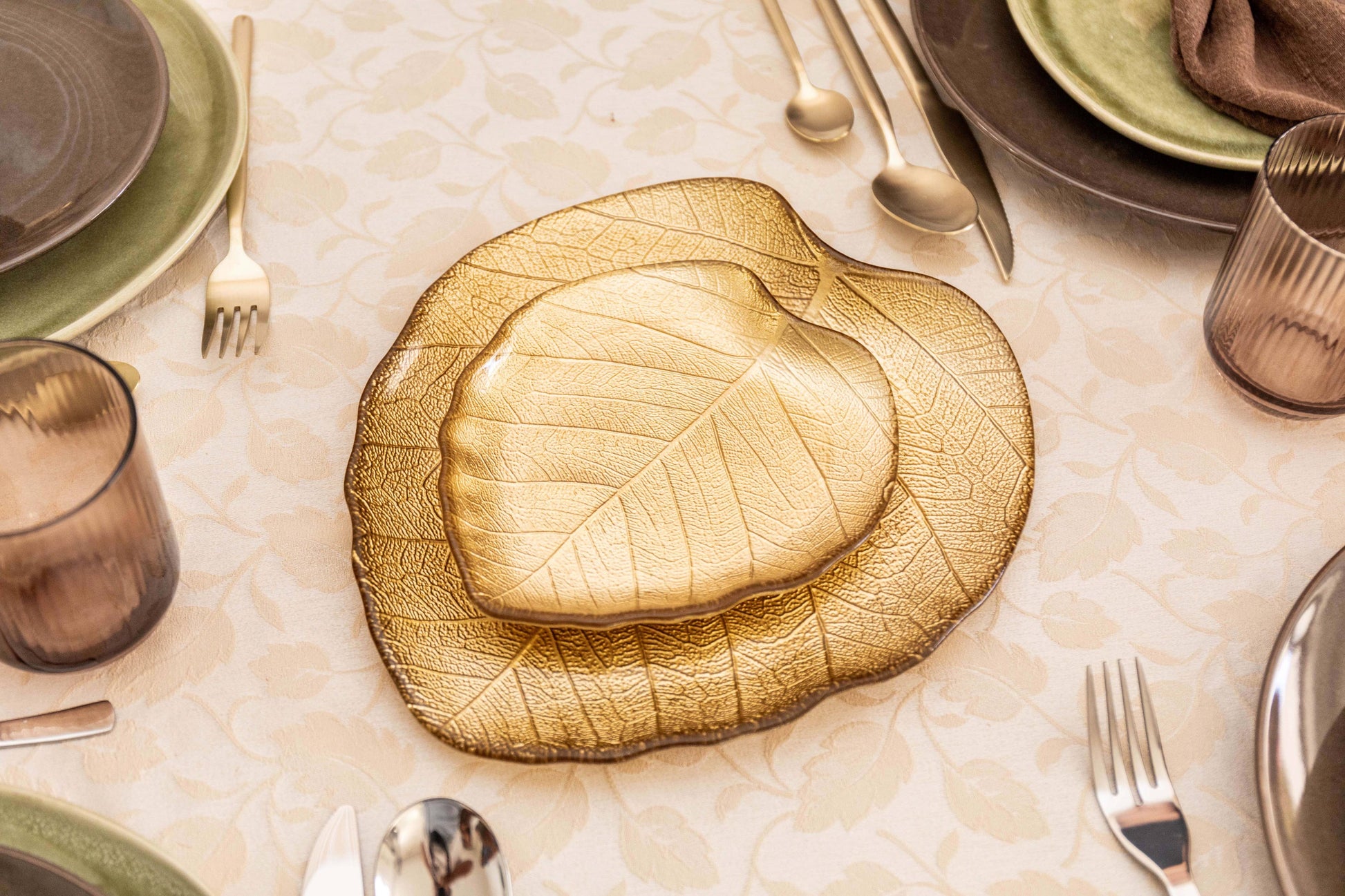 Auf einem gedeckten Tisch steht die AULICA - TASSE MIT GOLDENEM BLATT, GROSSES MODELL, 27X24X2,5 CM in der Mitte, umgeben von beigem und braunem Geschirr, goldenem Besteck und gerippten Gläsern auf einem cremefarbenen Tischtuch.