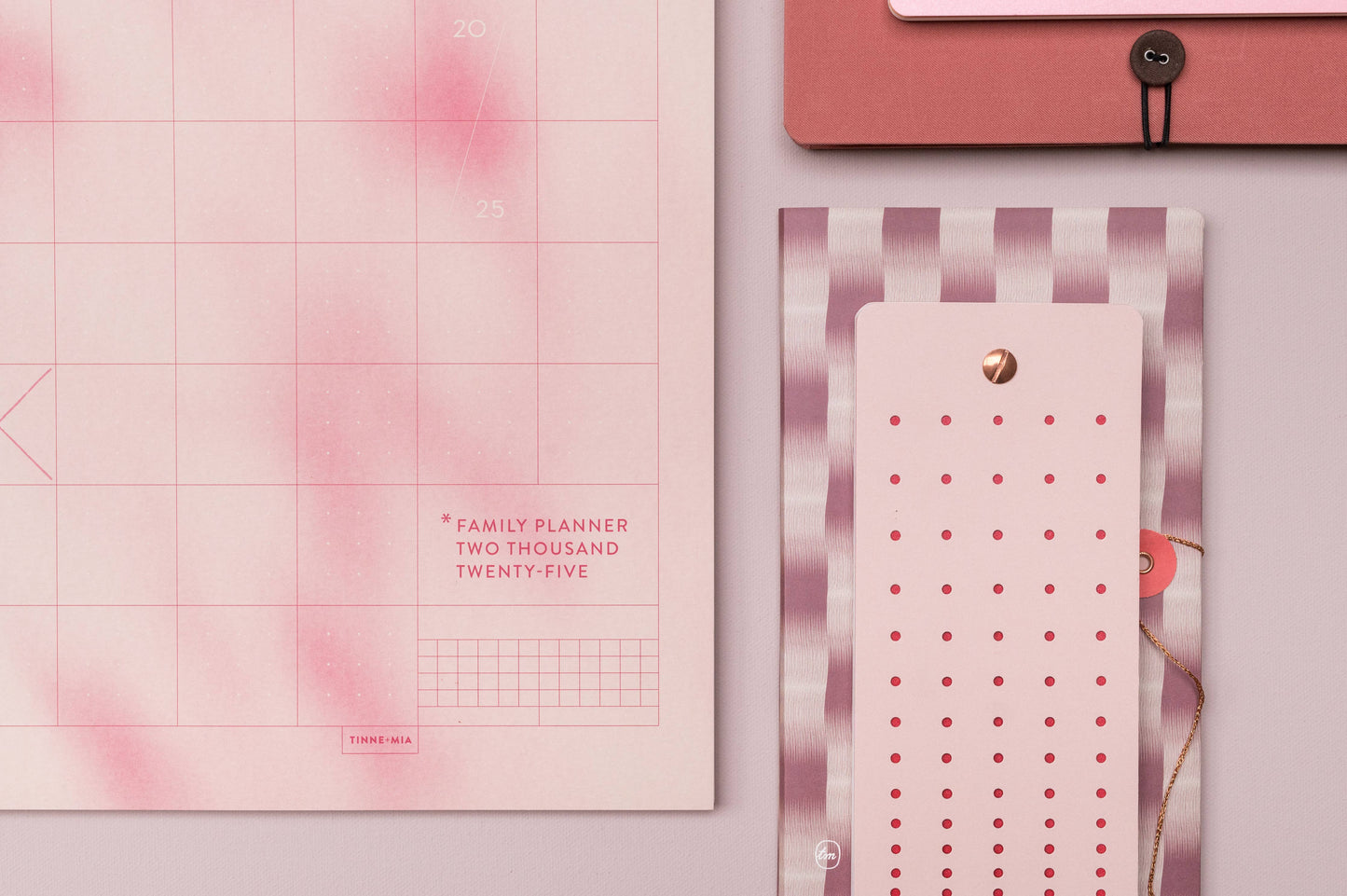 Eine Nahaufnahme einer rosafarbenen Wand mit dem Tinne Mia Aufgabenliste - Mellow Rose Familienplaner, einem gestreiften Notizblock aus FSC-zertifiziertem Papier und einem Teil eines rosafarbenen Umschlags mit schwarzem Knopfleistenverschluss.