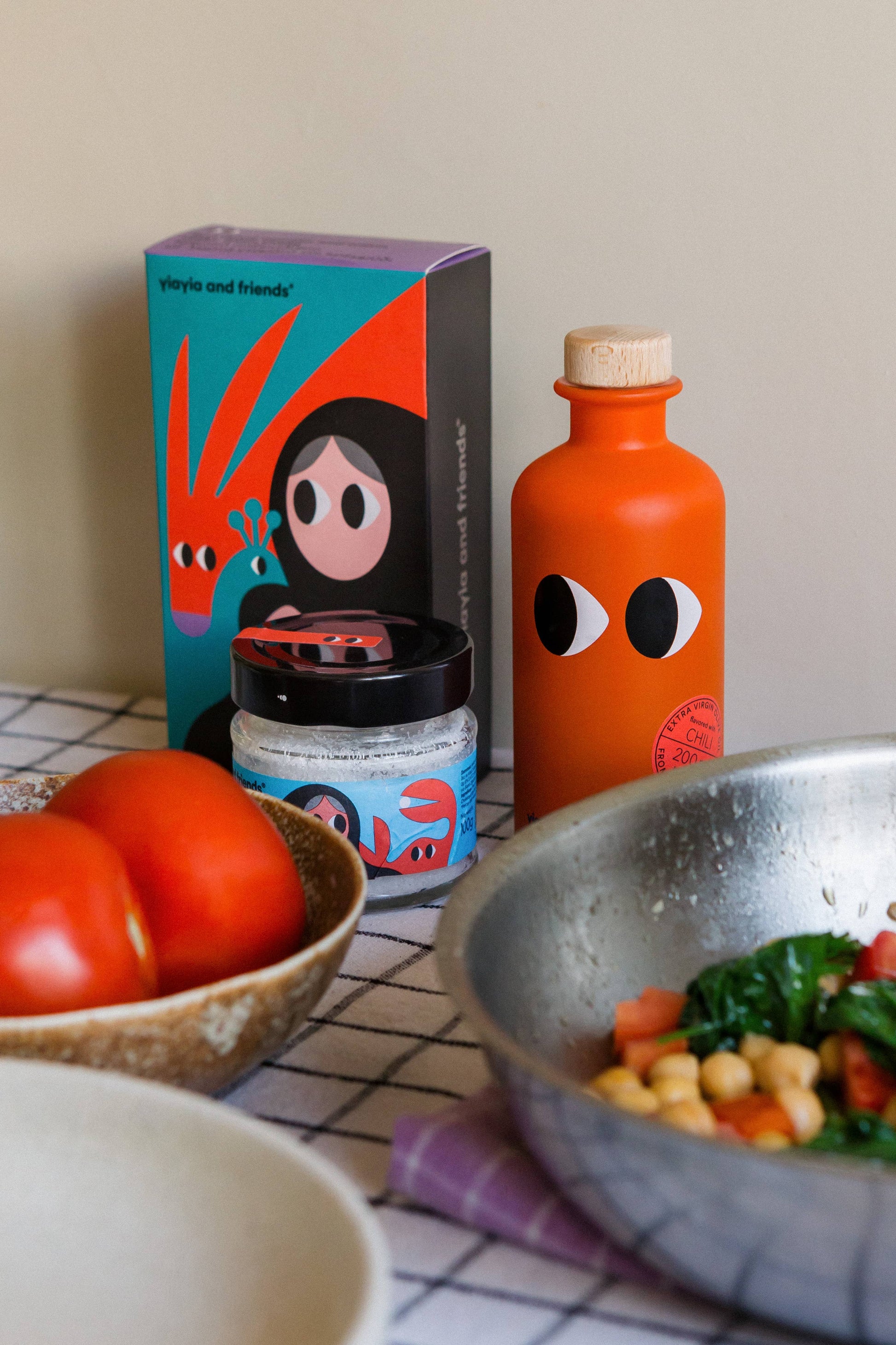 Eine lebhafte Küchenszene mit Tomaten in einer Schüssel, einer Pfanne mit Gemüse und Kichererbsen, die in yiayia and friends - Natives Olivenöl extra mit Chili gekocht wurden, sowie verspielte Gläser und Flaschen und auffällige grafische Schachteln von yiayia and friends.