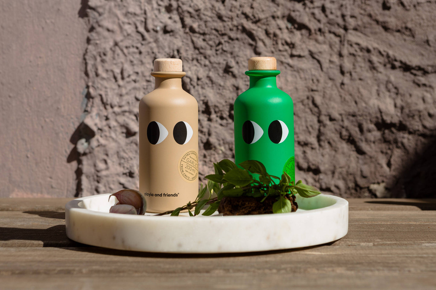 Zwei Flaschen von yiayia and friends - eine beige, eine grüne - stehen auf einem weißen Tablett zwischen Kräutern und Knoblauch. Verspielt und perfekt für ein Essen im Freien, enthalten sie yiayia and friends Natives Olivenöl Extra mit Knoblauch.