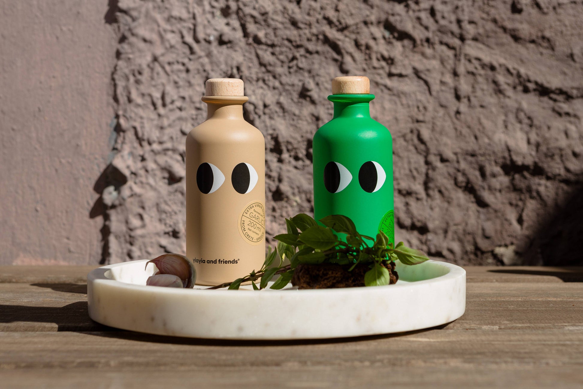 Zwei Flaschen von yiayia and friends - eine beige, eine grüne - stehen auf einem weißen Tablett zwischen Kräutern und Knoblauch. Verspielt und perfekt für ein Essen im Freien, enthalten sie yiayia and friends Natives Olivenöl Extra mit Knoblauch.