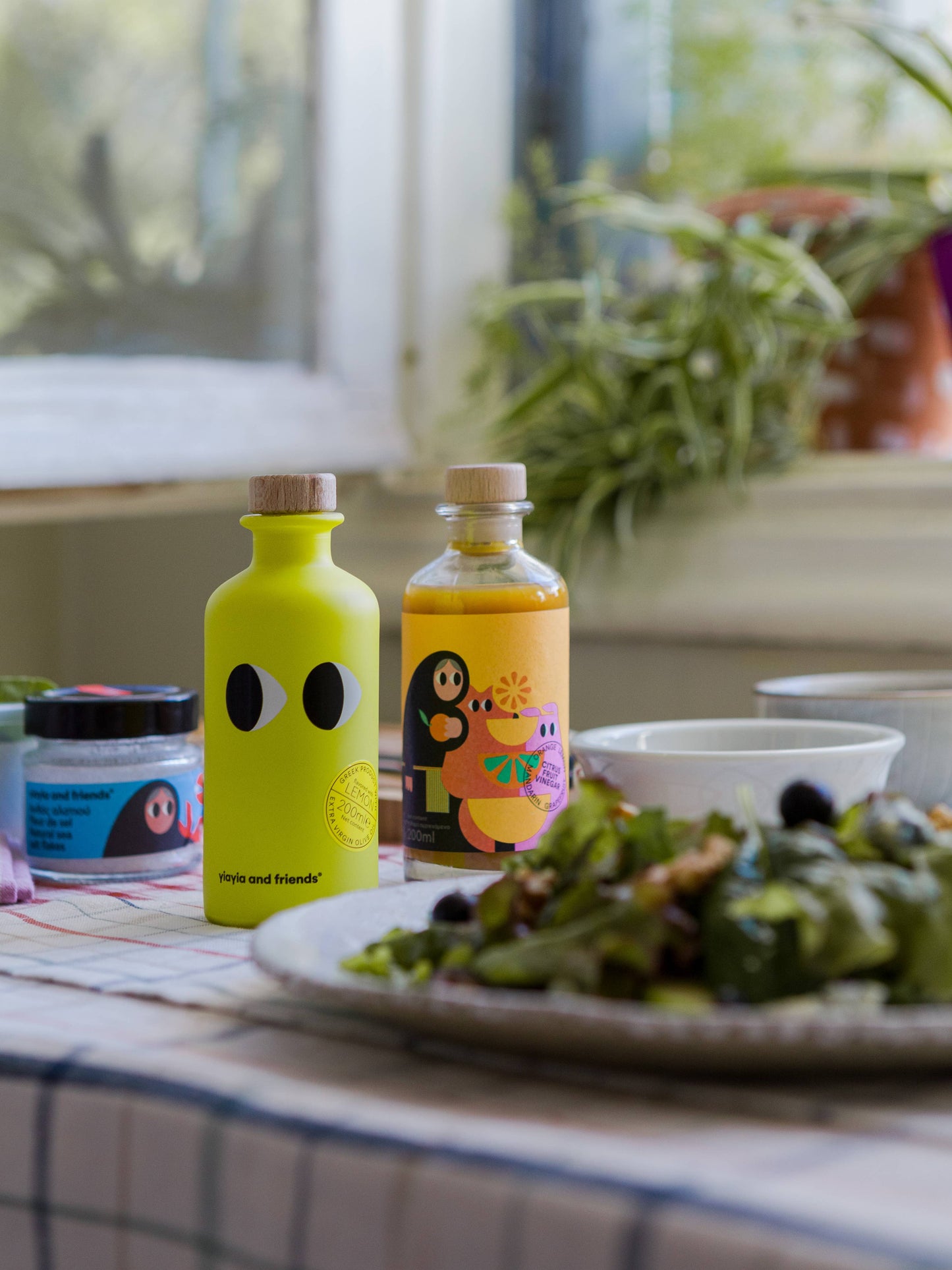Zwei leuchtende Flaschen - darunter yiayia and friends - Natives Olivenöl extra mit Zitrone - stehen auf einem karierten Tisch mit Salat, einer Schüssel und einem Glas. Ein Fenster und eine Topfpflanze schaffen im Hintergrund eine frische, gemütliche Küchenatmosphäre.