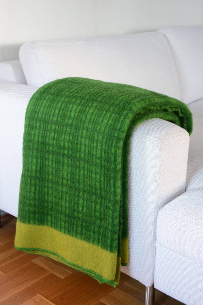 Die Darling Tagesdecke aus Lammwolle | Kiwi von Sofie Sjöström Design, in Grün mit hellgrünem Rand, liegt über dem Arm eines weißen Sofas; darunter ist ein Holzboden zu sehen.