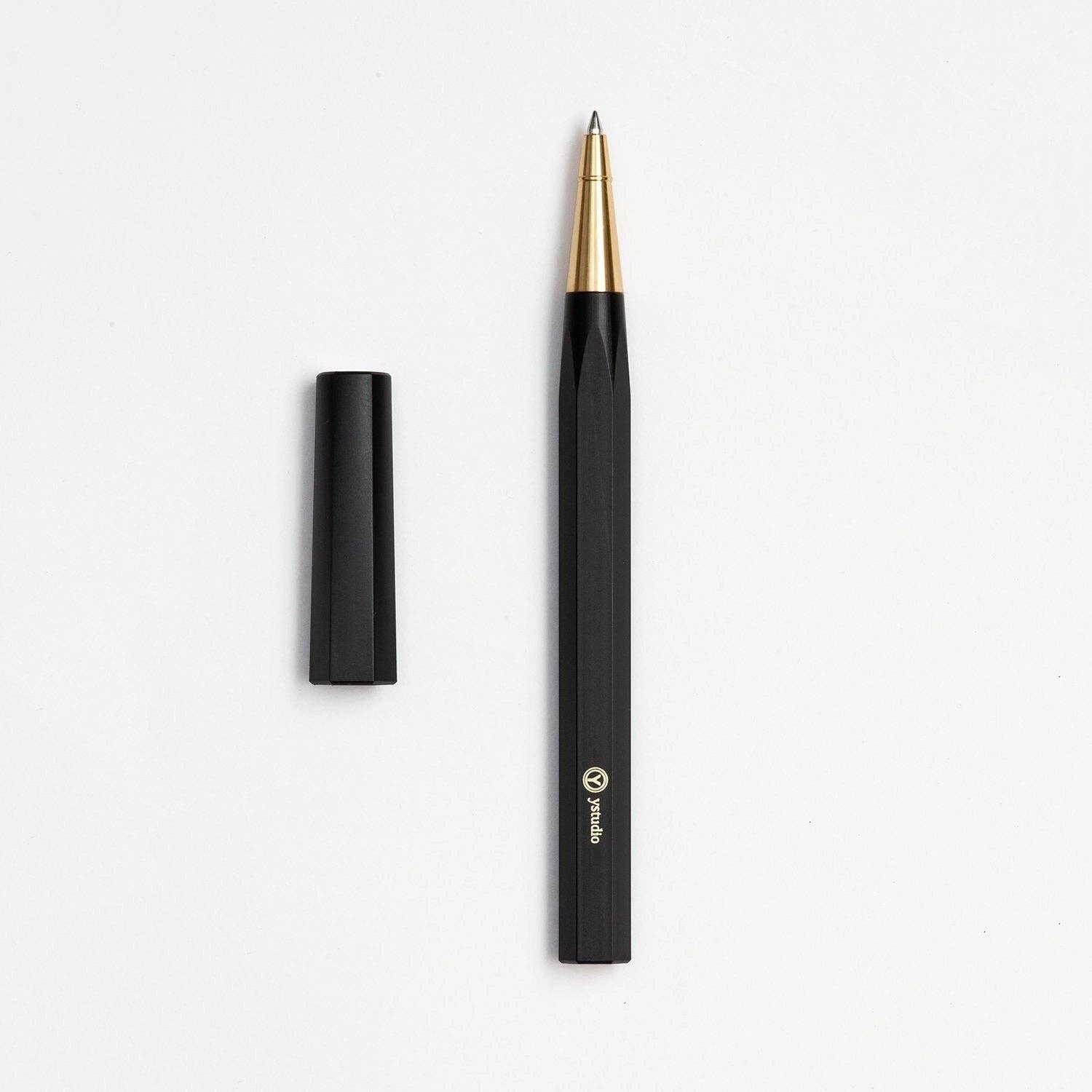 Der ENTREPÔT - YSTUDIO Harz-Rollerball-Stift (Schwarz) verfügt über eine goldene Spitze, eine schwarze Kappe auf der linken Seite und ein Markenlogo in der Nähe der Basis, alles vor einem weißen Hintergrund, um sein Schreibritual hervorzuheben.