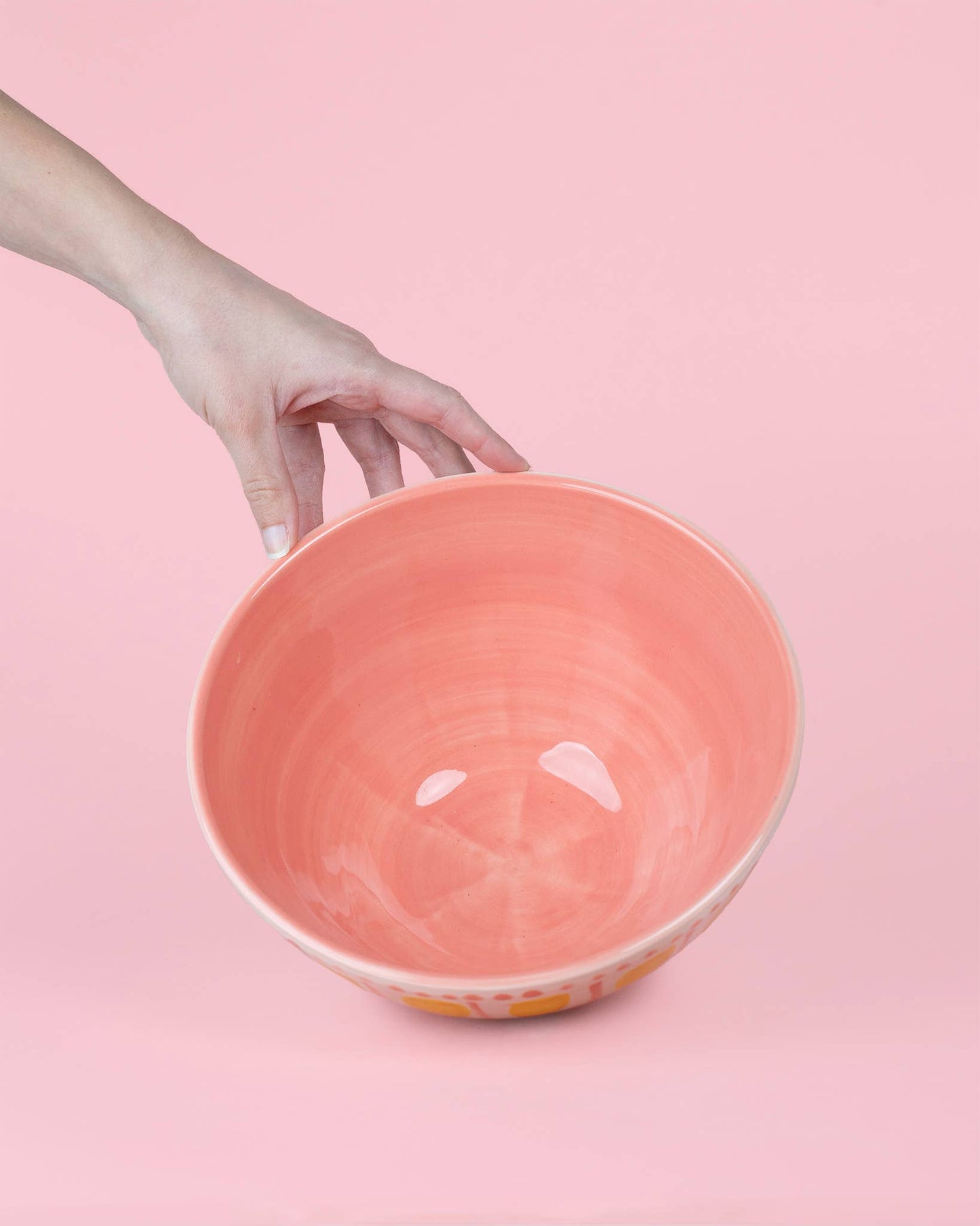 Eine Hand hält sanft die glänzende Alfar Tierra Cocida | Handgefertigte Keramik-Servierschale 1,5L Rosa und Gelb VALENTINA vor einem unifarbenen, blassrosa Hintergrund.