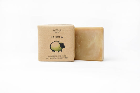 Lanola Seife von maisoap berlin: Naturseife mit Lanolin für trockene Haut, verpackt in Recyclingpapier mit einem Schaf und dem Text "LANOLA, handgerührte Seife mit natürlichen Zutaten" auf weißem Hintergrund.