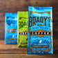 Drei farbenfrohe Tüten der Brady's Coffee Company sind in einer Reihe auf Holz angeordnet, wobei die vordere blaue Tüte - Brady's Signature Blend Kaffee - scharf abgebildet ist und die beiden anderen Spezialitätenkaffee-Tüten dahinter leicht verschwommen sind.
