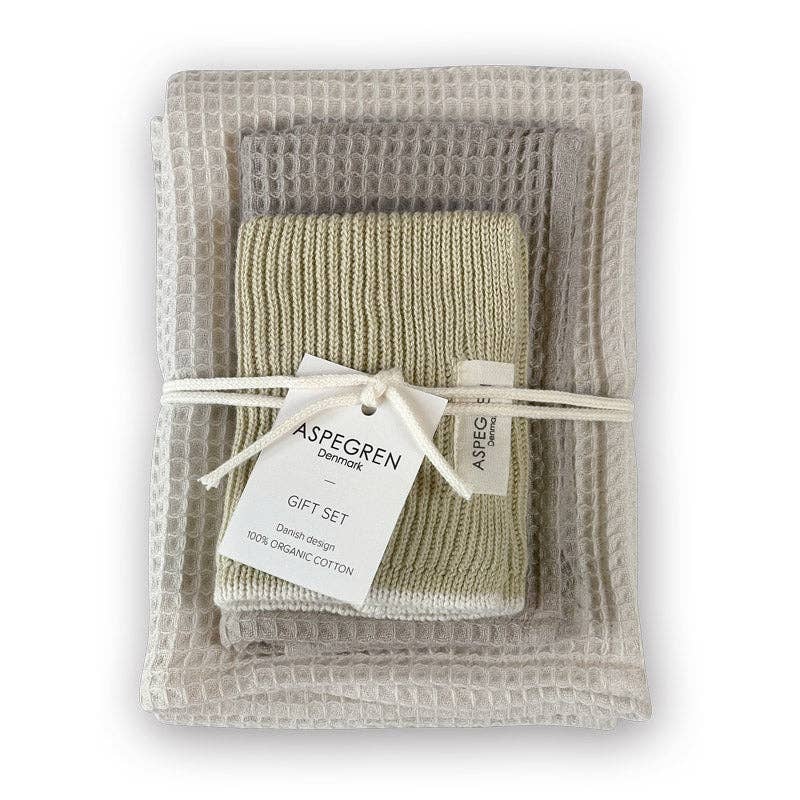 Ein Set gefalteter Waschlappen aus Bio-Baumwolle in neutralen Tönen, mit einer Schnur zusammengebunden und mit der Aufschrift „Aspegren Denmark – Geschenkset – Lemon Grass“ beschriftet, liegt auf einem strukturierten Handtuch mit Waffelmuster – ein umweltfreundliches Geschenk von Aspegren Denmark.