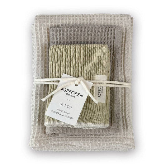 Ein Set gefalteter Waschlappen aus Bio-Baumwolle in neutralen Tönen, mit einer Schnur zusammengebunden und mit der Aufschrift „Aspegren Denmark – Geschenkset – Lemon Grass“ beschriftet, liegt auf einem strukturierten Handtuch mit Waffelmuster – ein umweltfreundliches Geschenk von Aspegren Denmark.