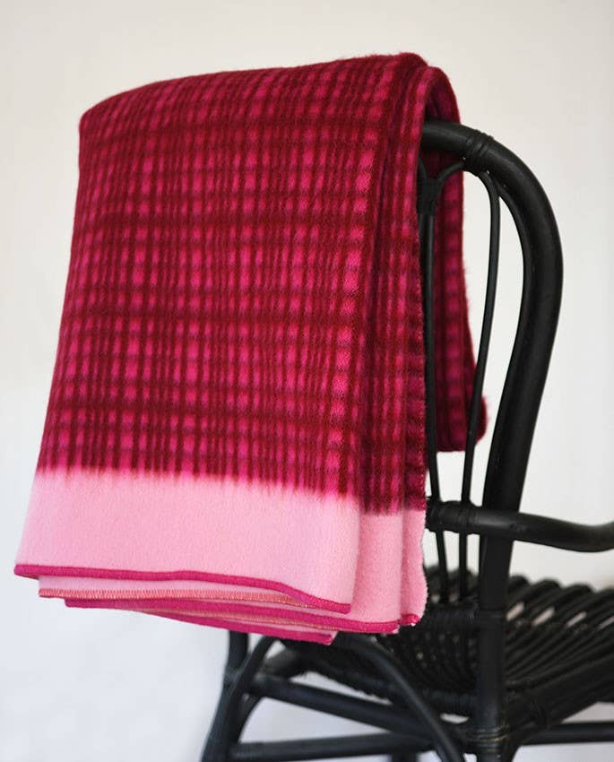 Eine gefaltete Sofie Sjöström Design Darling Tagesdecke aus Lammwolle in Fuchasia mit einem hellrosa Rand ist über die Rückenlehne eines schwarzen Korbstuhls drapiert.