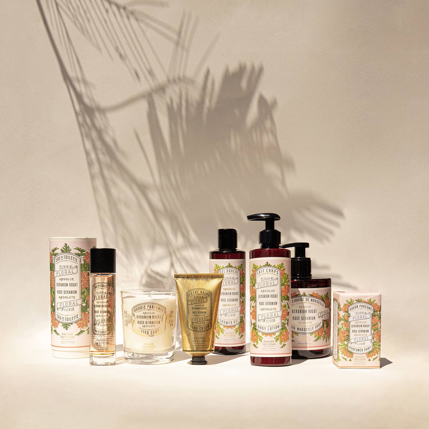 Eine Sammlung von Panier Des Sens-Produkten mit floralen Vintage-Etiketten, darunter Duftseife | Rosengeranie 150 g, eine Tube, eine Kerze und Schachteln - alle auf einer neutralen Fläche mit Palmblattschatten im Hintergrund angeordnet.