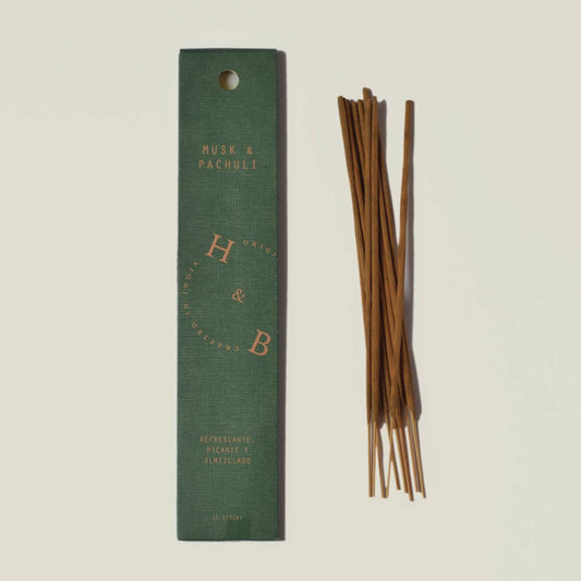Eine grüne H&B-Weihrauchdose mit der Aufschrift "Natürlicher Moschus- und Patchouli-Weihrauch" steht neben braunen Räucherstäbchen. Die Schachtel hat einen goldenen kreisförmigen Text, eine Bodenbeschreibung und erinnert an Noten von natürlichem Moschus und Patchouli.