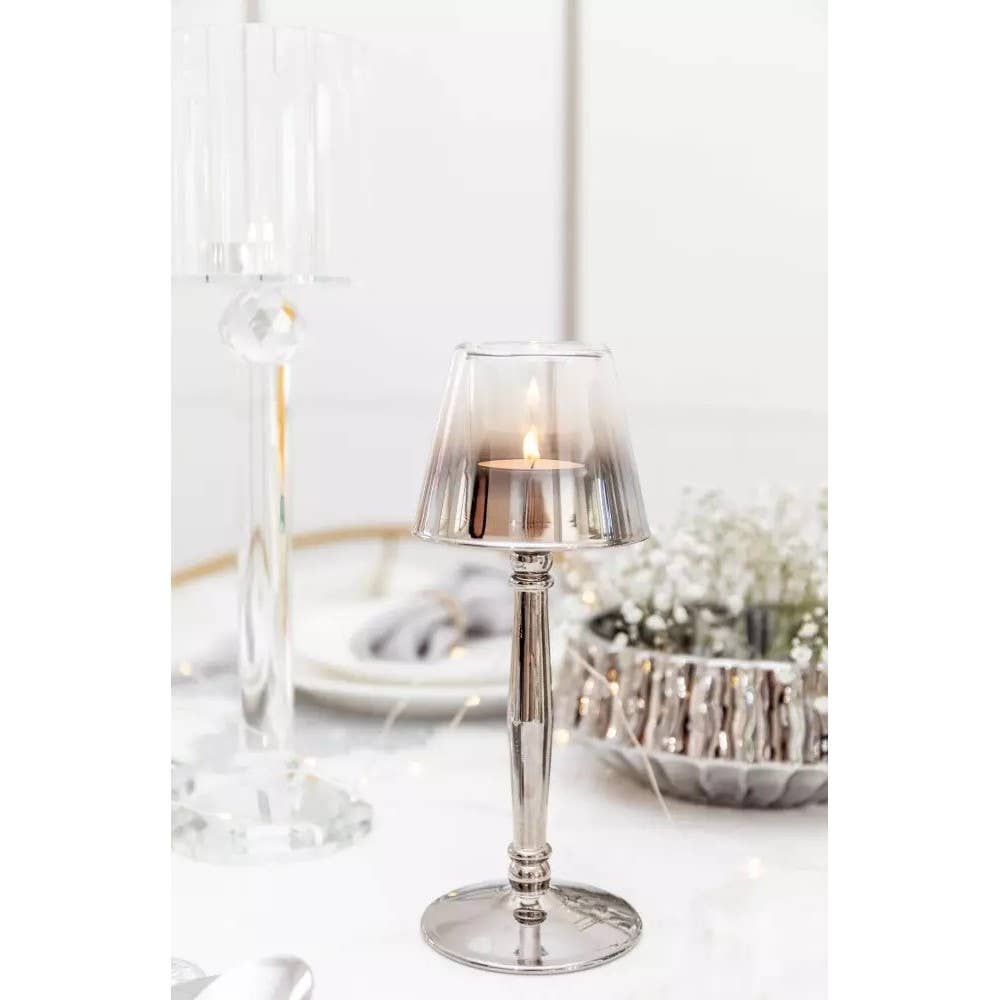 Der Silberne Kerzenhalter auf Fuß (9,5x9,5x22,5cm) von AULICA steht auf einem eleganten weißen Tisch mit dekorativen Wohnaccessoires, Tellern und einer Schale mit weißen Blumen für ein helles, anspruchsvolles Ambiente.