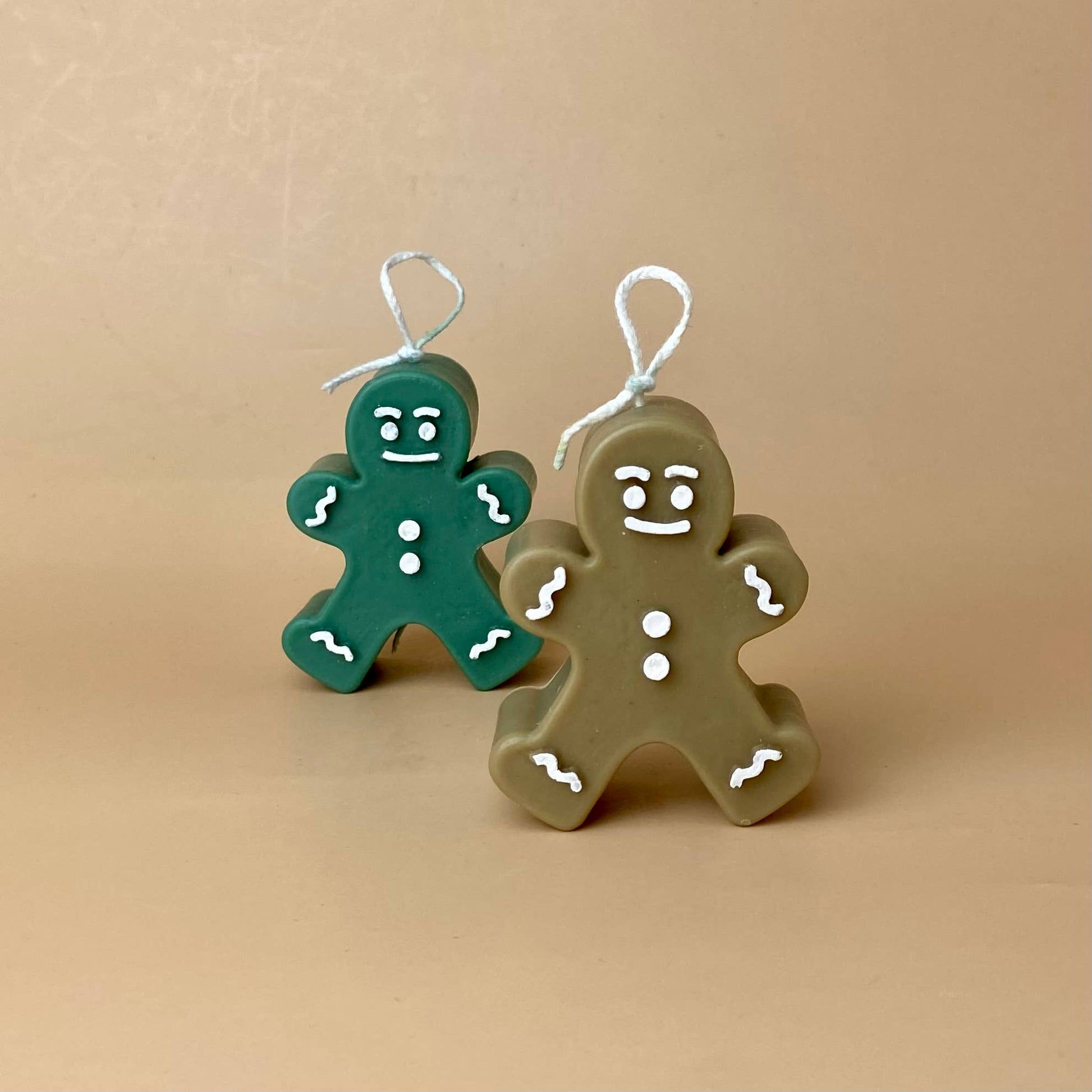 Zwei SOULCandle - Weihnachtskerzen von SOULCandle in Form von Lebkuchenornamenten – eine grün und eine braun – verfügen über weiße Zuckergussdetails und Aufhängeösen und werden aufrecht vor einem beigen Hintergrund gezeigt.