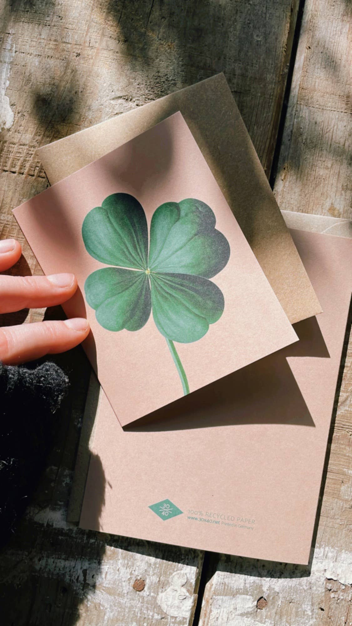 Eine Hand hält die 30x40 Mini-Karte "Paul [Kleeblatt]", gedruckt in Deutschland auf Recyclingpapier, auf einem braunen Umschlag auf rustikalem Holz. Sonnenlicht wirft weiche Schatten über die Szene.