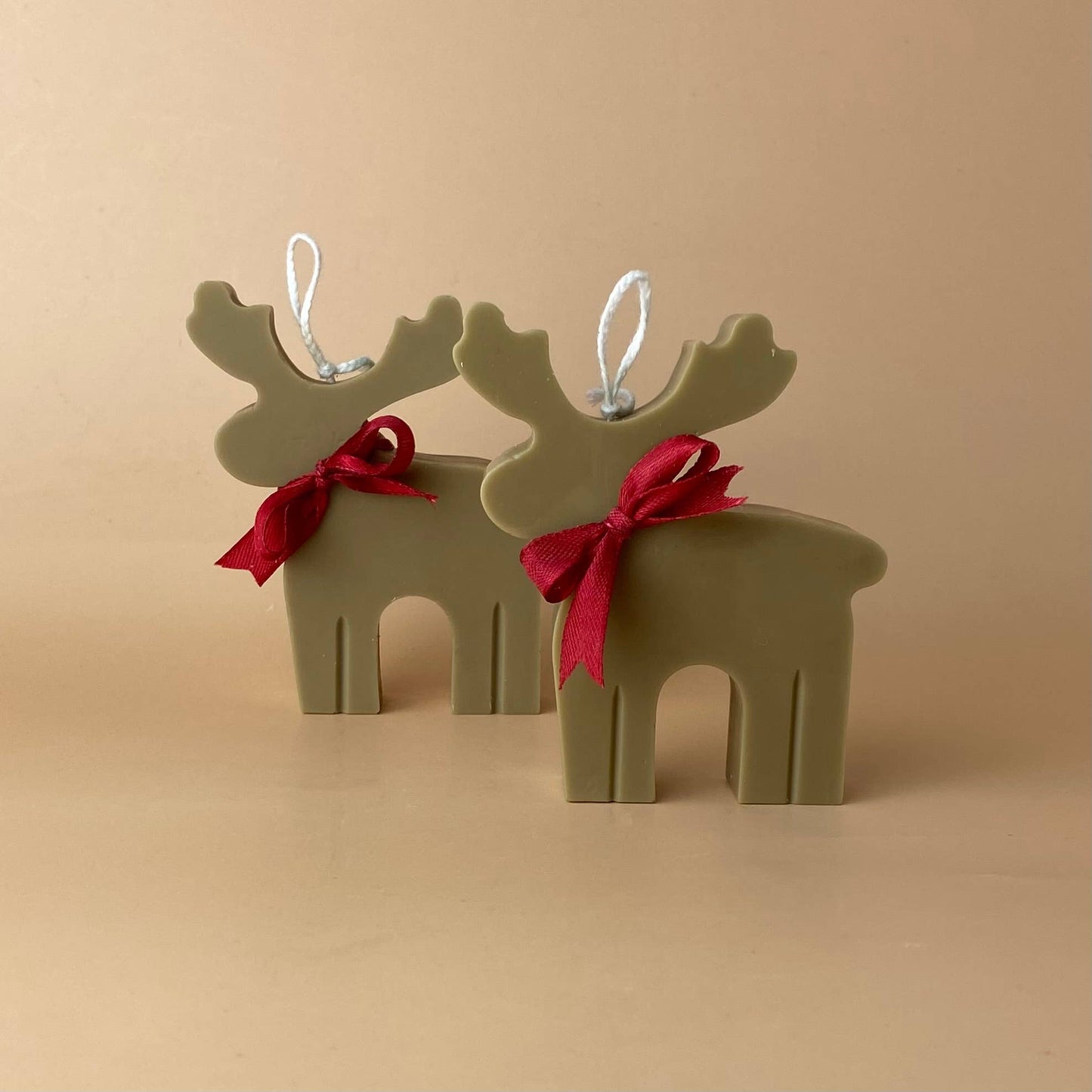 Zwei SOULCandle - Weihnachtskerzen in Rentierform, handgefertigt in Olivgrün mit roten Schleifen und weißen Aufhängeösen, werden auf einer beigen Fläche mit passendem beigen Hintergrund präsentiert.