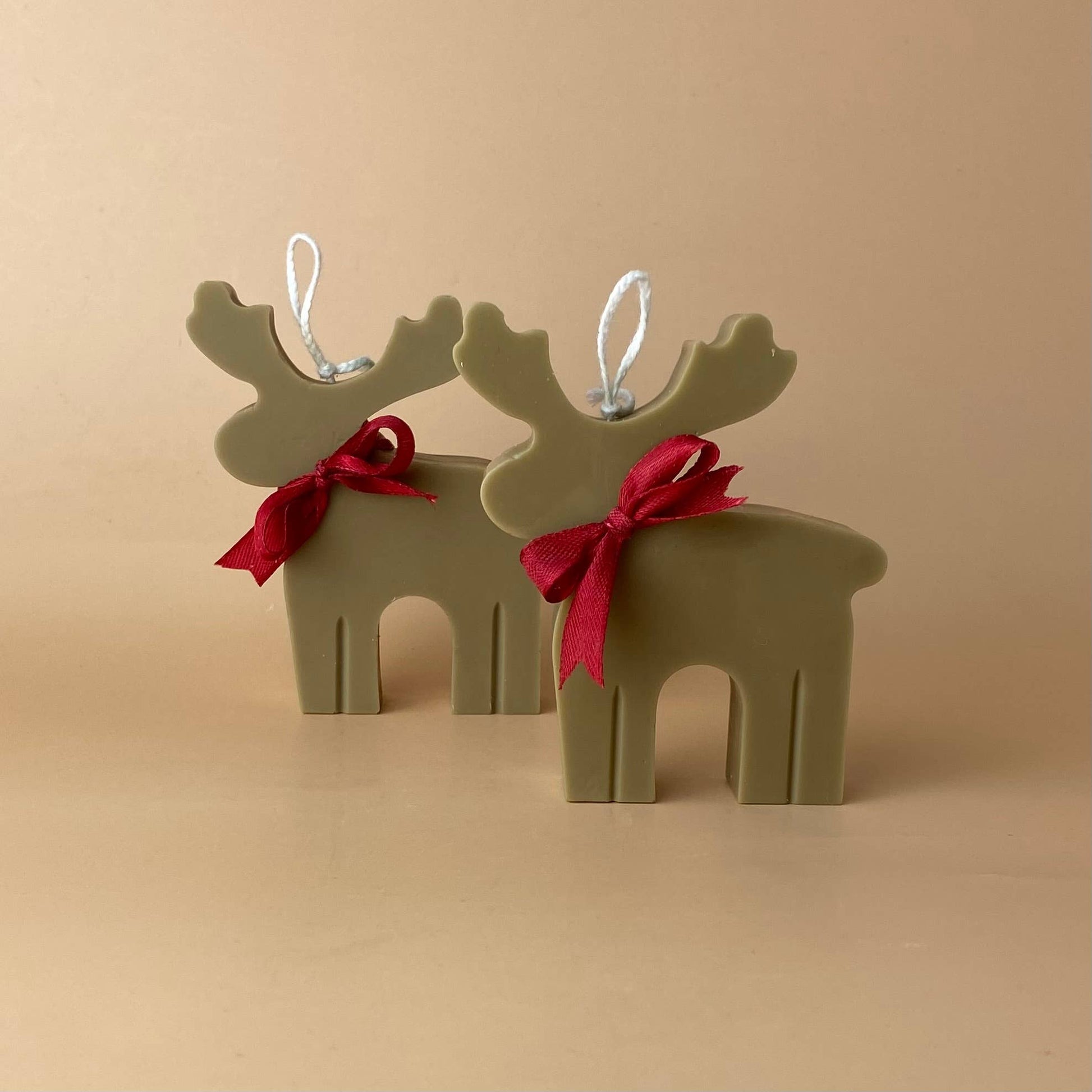 Zwei SOULCandle - Weihnachtskerzen in Rentierform, handgefertigt in Olivgrün mit roten Schleifen und weißen Aufhängeösen, werden auf einer beigen Fläche mit passendem beigen Hintergrund präsentiert.