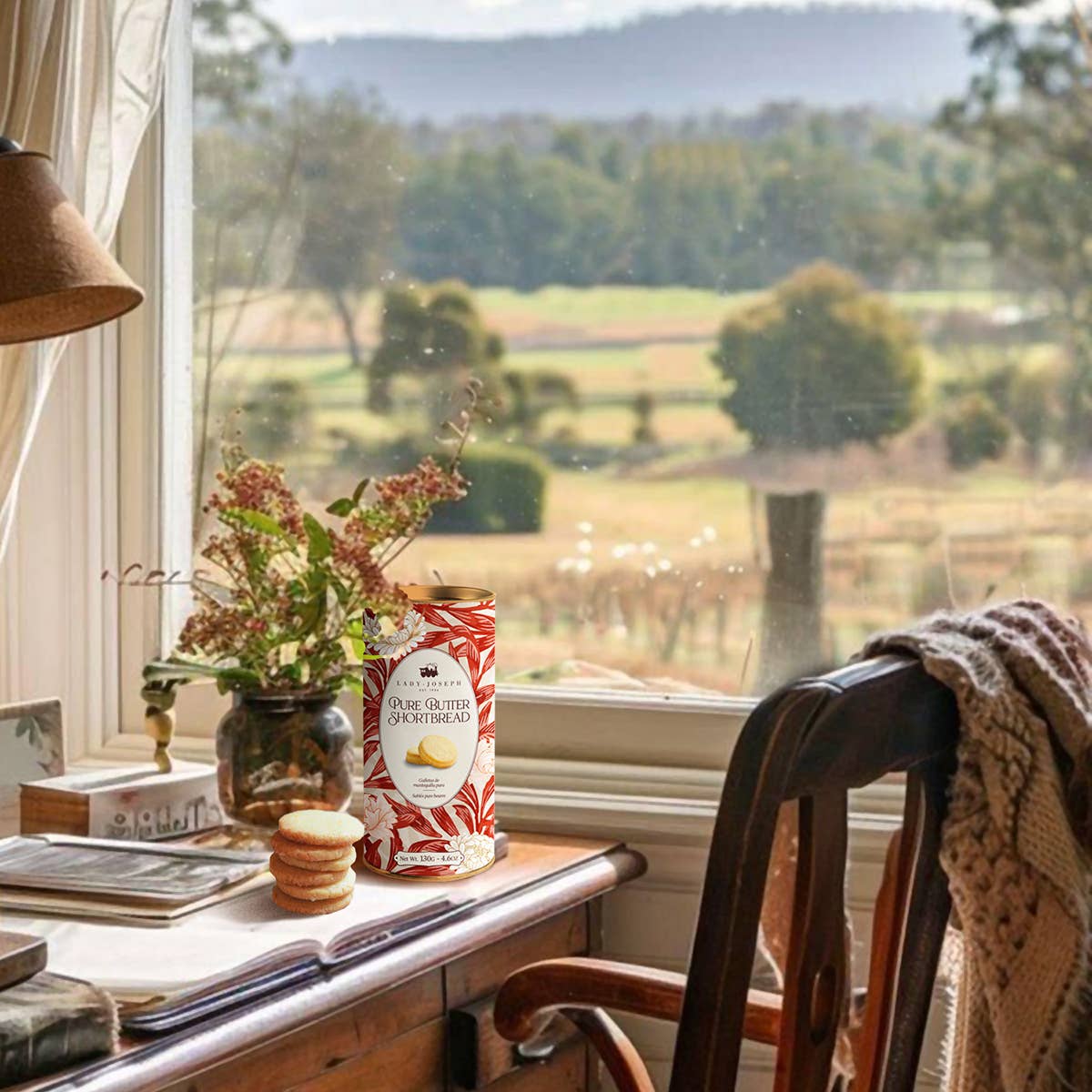 Ein gemütlicher Schreibtisch an einem Fenster mit Blick auf die Landschaft. Darauf sitzt Lady Joseph Buttergebäck aus reiner Butter Doppel-Butter-Shortbread 130g, eine Vase mit Blumen, eine Lampe, Bücher und eine Strickdecke, die über den Holzstuhl drapiert ist.