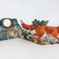 Die Fuchs Collage Karte von Mark Hearld by Art Angels zeigt einen leuchtend roten Fuchs mit gelben Augen in einem Wald unter einem sternenklaren Nachthimmel, umgeben von Pflanzen, Kaninchen und einem Vollmond im unverkennbaren Stil von Mark Hearld.