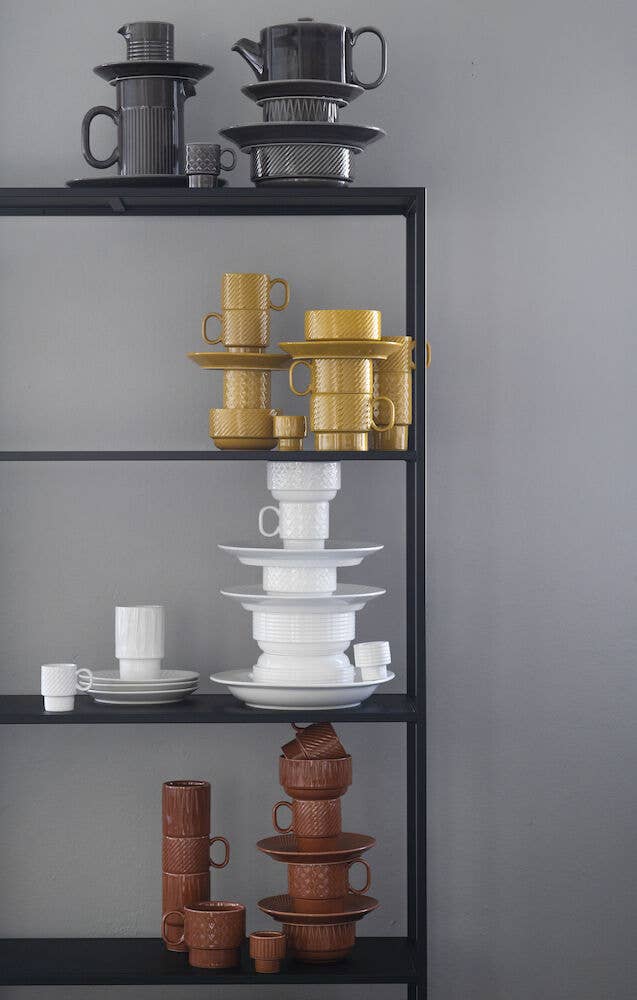 Auf einem schwarzen Metallregal sind vor einer grauen Wand ordentlich angeordnete zweiteilige Kaffeebecher von Sagaform GmbH Coffee & More in Schwarz, Gelb, Weiß und Braun sowie passende Tassen, Teekannen und Untertassen ausgestellt.