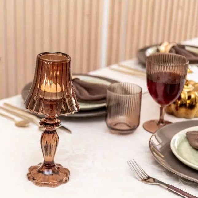 Ein gedeckter Tisch mit AULICA Rippengläsern, beigen Tellern, goldenem Besteck und dem AULICA - Brauner Kerzenhalter auf Glasfuß 10x10x24,5cm als Mittelstück auf einer weißen Tischdecke.