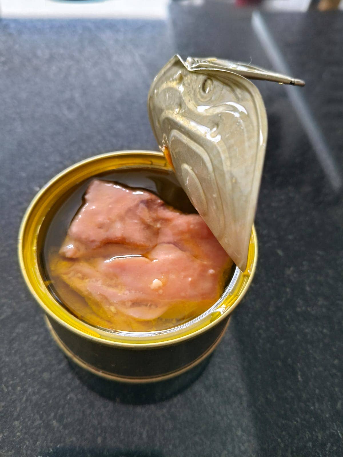 Eine geöffnete Dose Sardinha - Geräucherter Bonito-Thunfisch aus den Azoren in Olivenöl liegt auf einer dunklen Fläche, ihr Deckel ist teilweise aufgeschlagen und gibt den Blick auf rosa Scheiben geräucherten Thunfischs in Olivenöl frei. Marke: Sardinha.