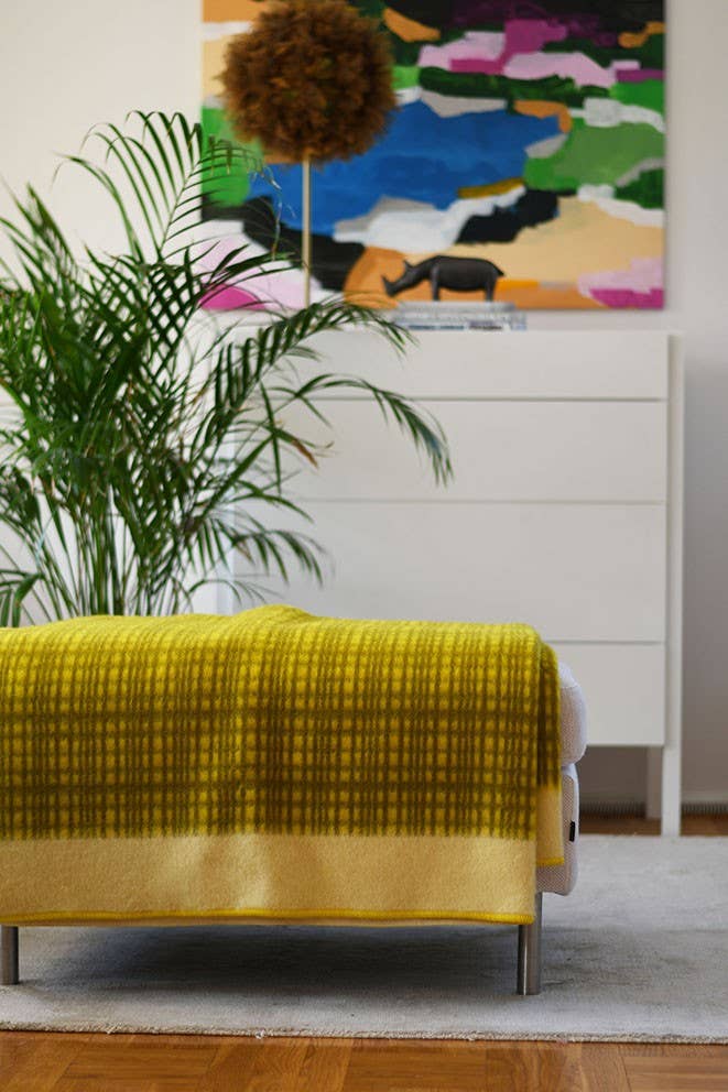 Ein modernes Wohnzimmer mit der gelben Darling Tagesdecke aus Lammwolle von Sofie Sjöström Design auf einer Bank, einer Blattpflanze, einer weißen Kommode und einem bunten abstrakten Gemälde im Hintergrund.