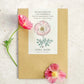 Ein Saatgutpäckchen Jora Dahl | Eschscholzia californica 'Thai Silk Appleblossom' (Goldmohn) mit Blumenillustration liegt hell platziert neben einem kleinen Stängel und zwei rosa Mohnblüten.