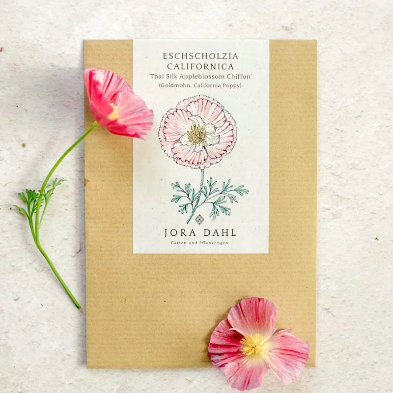 Ein Saatgutpäckchen Jora Dahl | Eschscholzia californica 'Thai Silk Appleblossom' (Goldmohn) mit Blumenillustration liegt hell platziert neben einem kleinen Stängel und zwei rosa Mohnblüten.