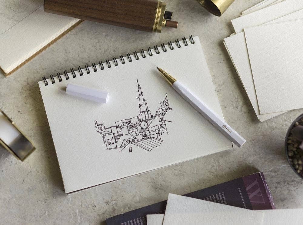 Ein spiralgebundenes Skizzenbuch zeigt eine Federzeichnung von Dächern und einem hohen Kirchturm. In der Nähe befindet sich der ENTREPÔT - YSTUDIO - Harz-Rollerball-Stift (Weiß), ein Radiergummi, Blankokarten, ein Buch und ein Federmäppchen auf einer hellen Fläche.