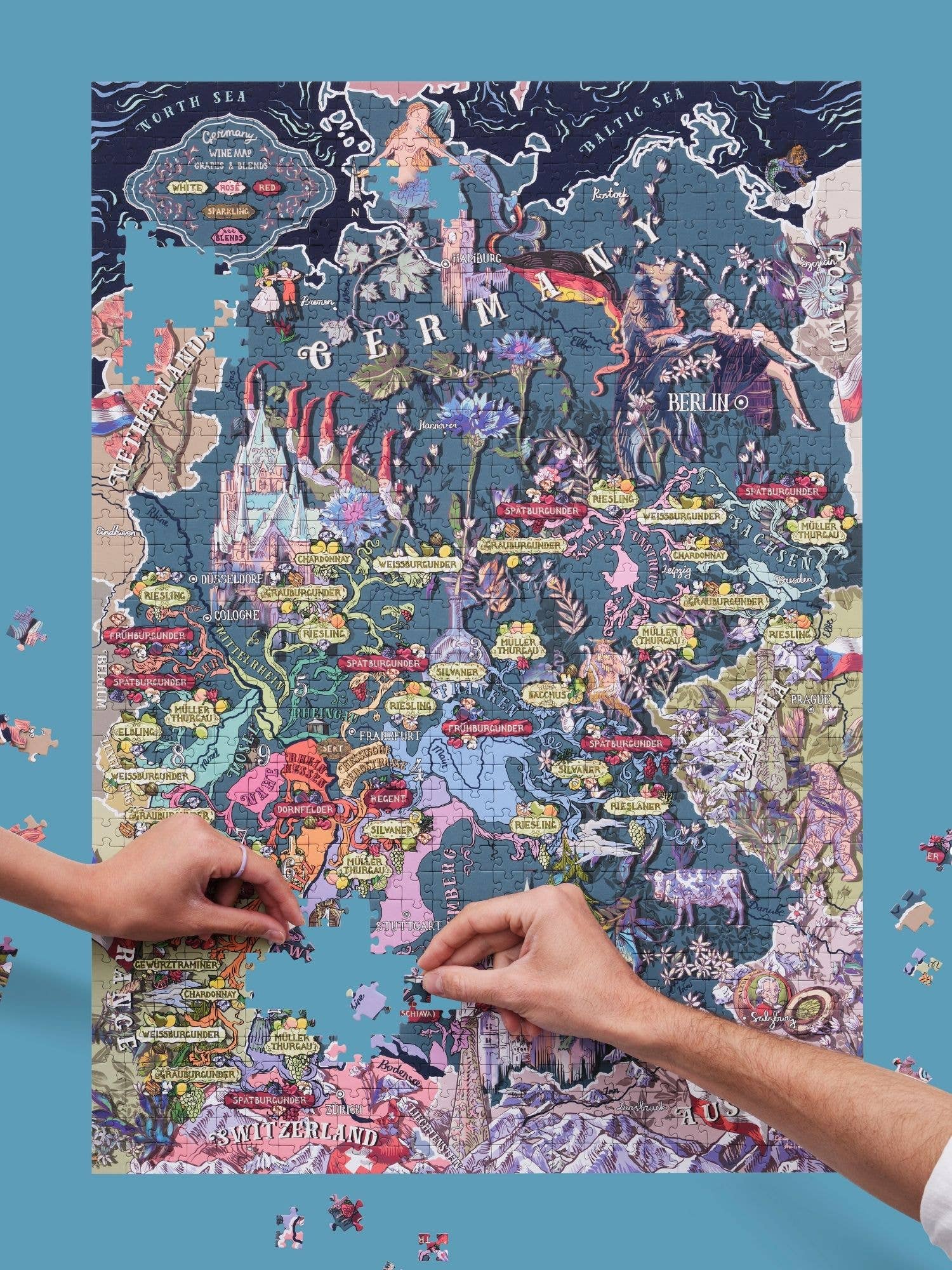Zwei Personen setzen das Water & Wines - Wein Puzzle - Deutschland von Water & Wines zusammen, ein leuchtend blaues Puzzle, das Deutschlands Regionen, Wahrzeichen, Tiere und berühmte Weinregionen mit bunten Illustrationen und stilisiertem Text zeigt.