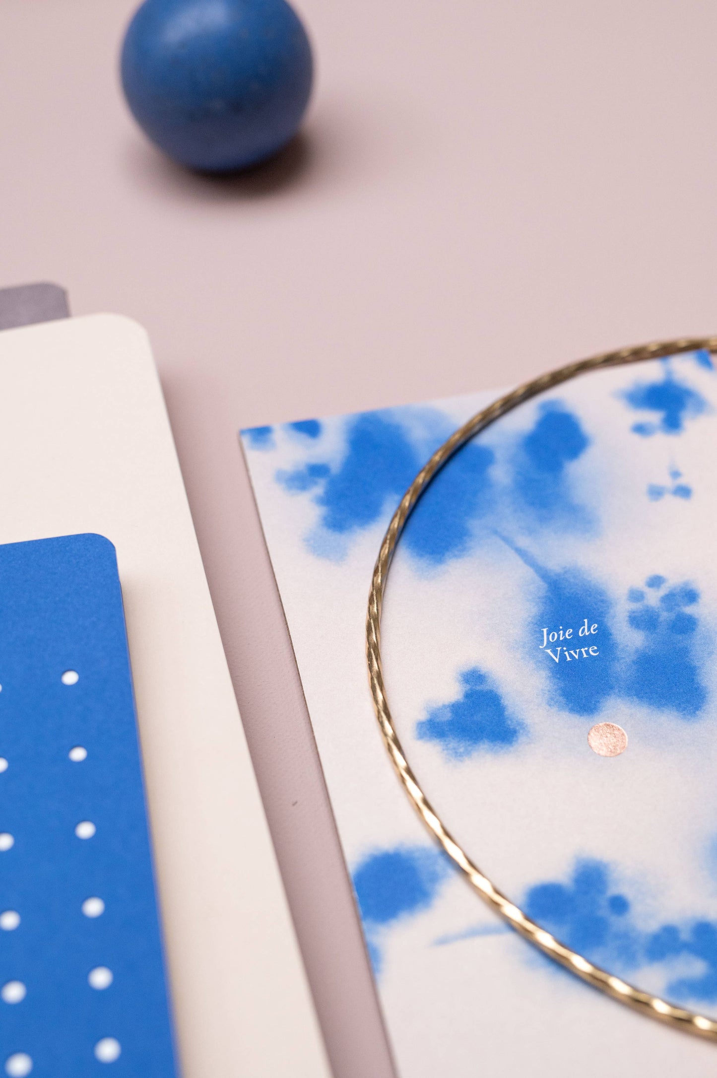 Blaue und weiße Papeterie mit der Tinne Mia - Aufgabenliste - True Blue auf FSC-Papier, einer Joie de Vivre-Karte mit blauen Aquarelltupfen, einem gedrehten Goldring und einer blauen Kugel auf hellviolettem Untergrund. Nachhaltig produziert.