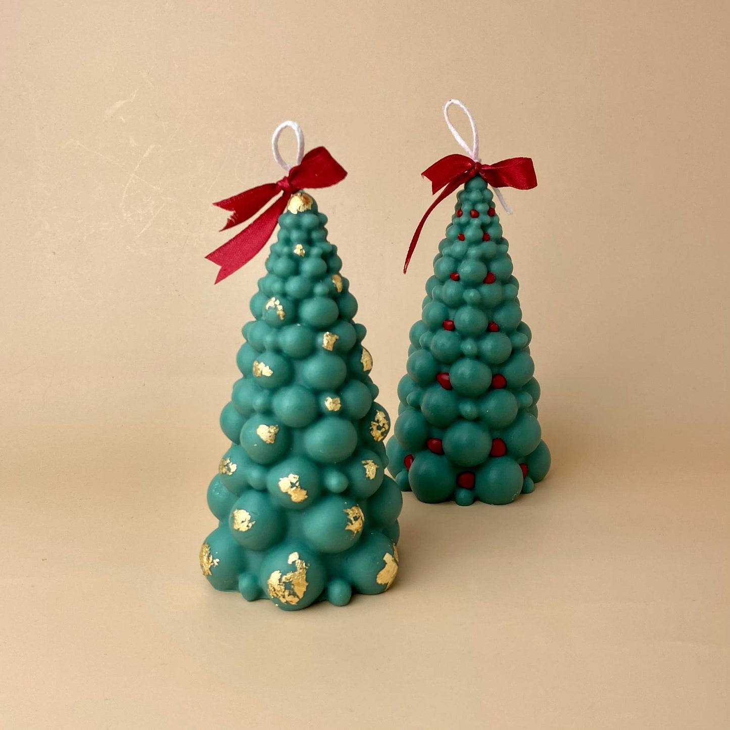 SOULCandle - Weihnachtskerzen von SOULCandle: Zwei handgefertigte grüne Christbaumkerzen aus Wachs mit weißem Docht, roten Schleifen und runden Ornamenten – eine mit goldenen Akzenten, die andere mit roten Punkten. Präsentiert auf schlichtem beigem Hintergrund.