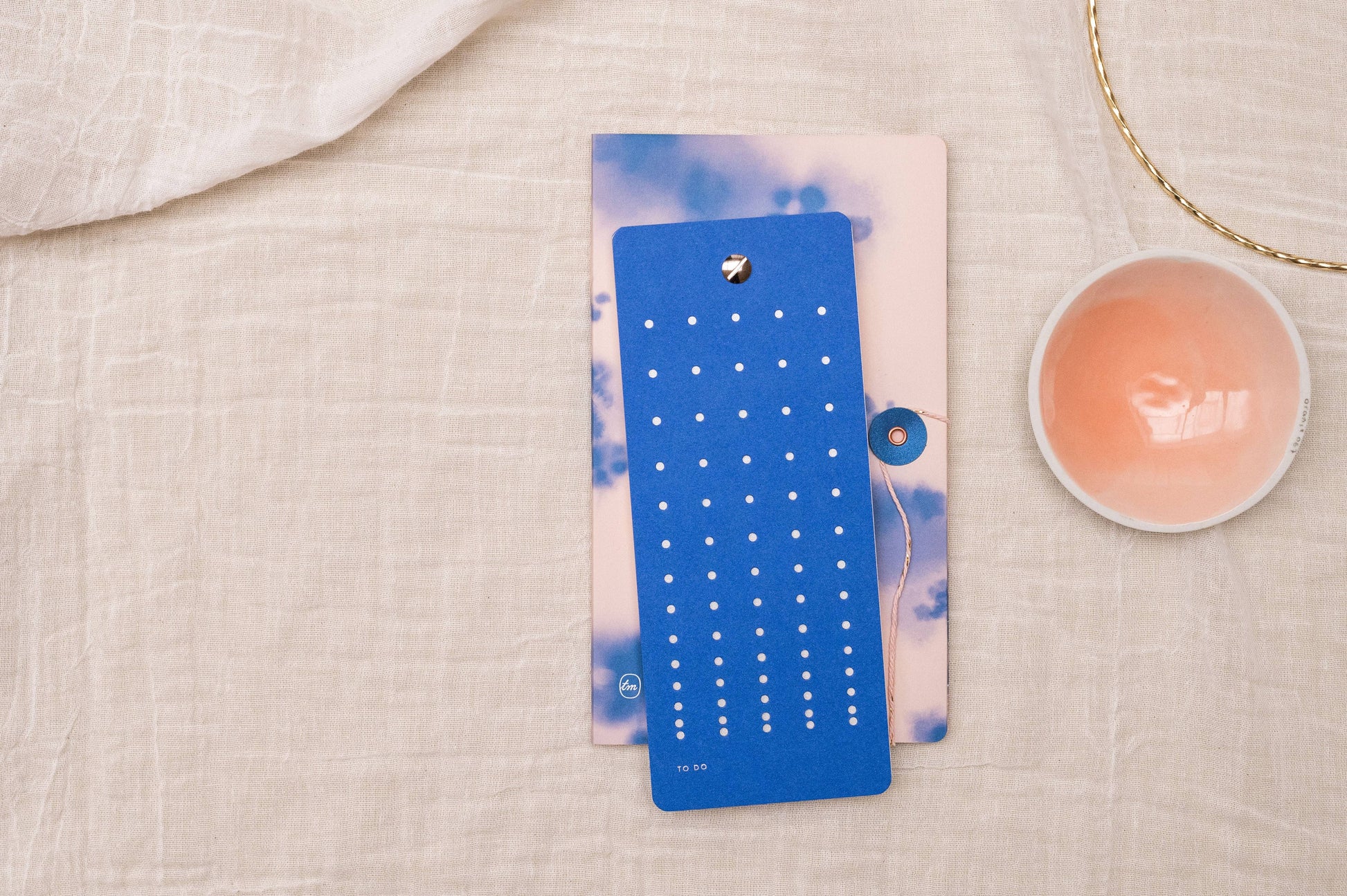 Die "Tinne Mia - Aufgabenliste - True Blue" aus FSC-Papier liegt auf einem farbigen Notizbuch neben einer rosa Keramikschale und einem goldenen Armband - nachhaltig produziert von Tinne Mia für umweltbewusstes Planen.