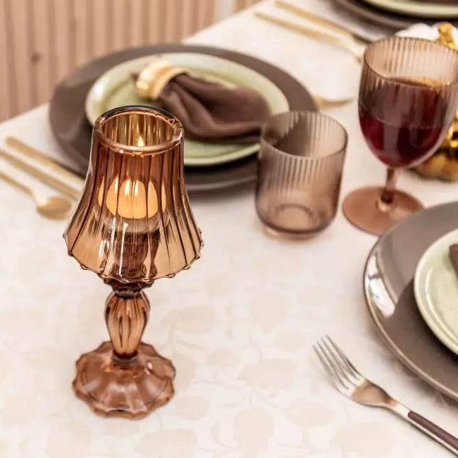 Ein für eine Mahlzeit gedeckter Tisch mit AULICA Goldbesteck, grünen Tellern, getönten Gläsern und einem AULICA Brauner Kerzenhalter auf Glasfuß (10x10x24,5cm), der eine bernsteinfarbene Kerze in der Mitte hält.