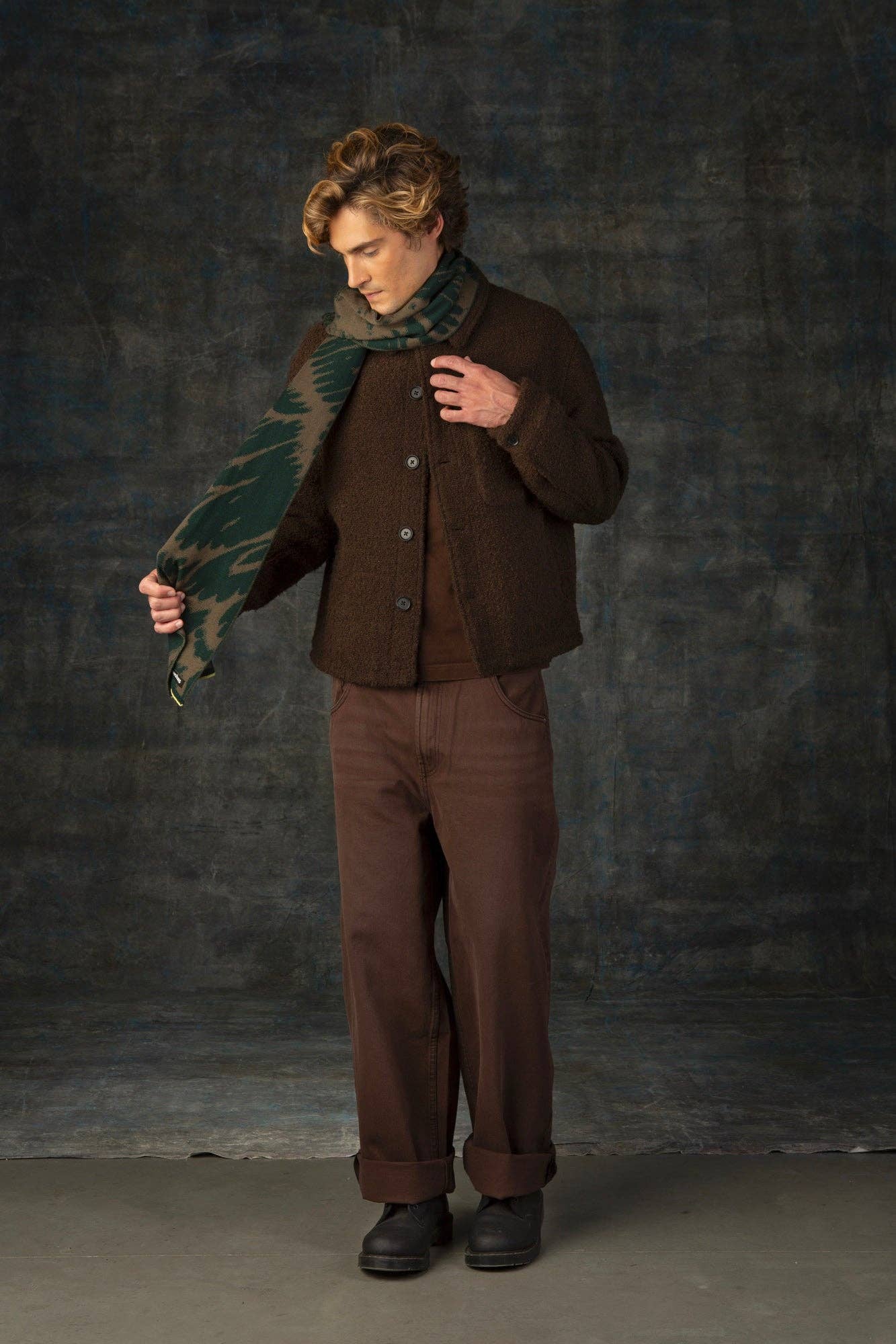 Eine Person mit gewelltem Haar blickt nach unten, während sie den Wolvis Schal aus Merinowolle - Furcula Wald Midi einstellt. Sie ist mit einer braunen Jacke, einer weiten braunen Hose und schwarzen Schuhen bekleidet und steht vor einem dunkel strukturierten Hintergrund.