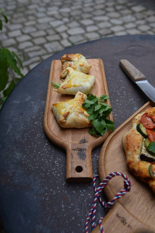 Drei goldbraune Gebäckstücke mit frischer Petersilie ruhen auf der LE RÉGAL - Kleine Schaufel, einem Aperitifbrett aus französischem Eichenholz von LE RÉGAL. Das Holzbrett steht im Freien neben einer Gemüsetarte, im Hintergrund sind Pflastersteine zu sehen.