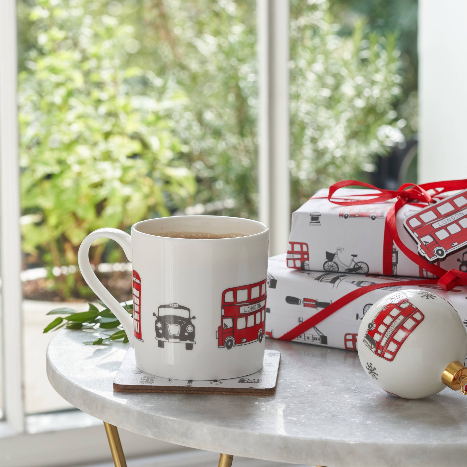 Der Victoria Eggs - London Skyline Becher, ein weißer Becher aus feinem Knochenporzellan mit roten Bus- und Taximotiven, steht auf einem Untersetzer neben einer passenden Christbaumkugel und Souvenirs auf einem runden Marmortisch neben einem sonnendurchfluteten Fenster mit viel Grün draußen.