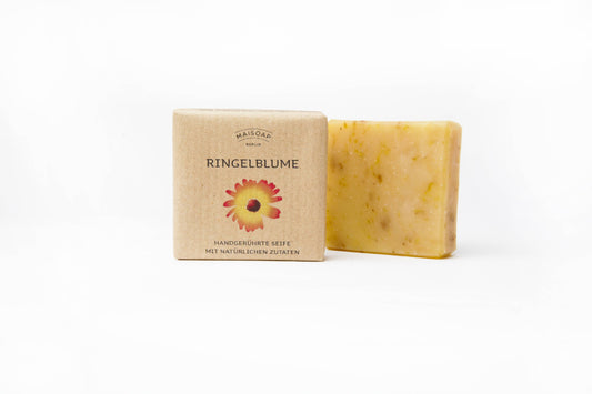 Ein Stück maisoap berlin - Ringelblume Seife liegt neben der Box mit Calendula-Blüten-Illustration - eine natürliche, sanft pflegende Seife der Marke maisoap berlin, bekannt für unterstützende Wirkung bei Wundheilung.