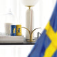 Eine Nahaufnahme des Tisches zeigt eine schwedische Flagge, Solstickan Design Streichholzröhrchen - Gelb/Blau (nur EU), eine weiße Tasse, eine Glaskuppel, einen Stapel Bücher, eine weiße Lampe und einen Kerzenhalter aus Messing mit einer rosa Kerze.